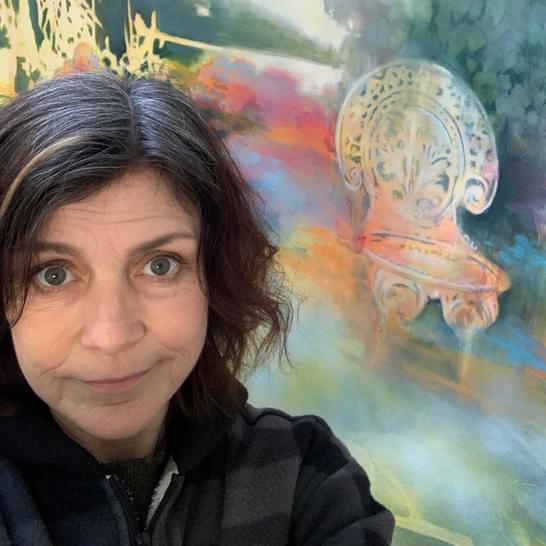Angela is next to a colourful painting, with long black hair wearing a black coat