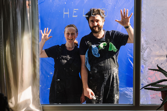 Two adults wave through a window with the word 'hello' written on the window.