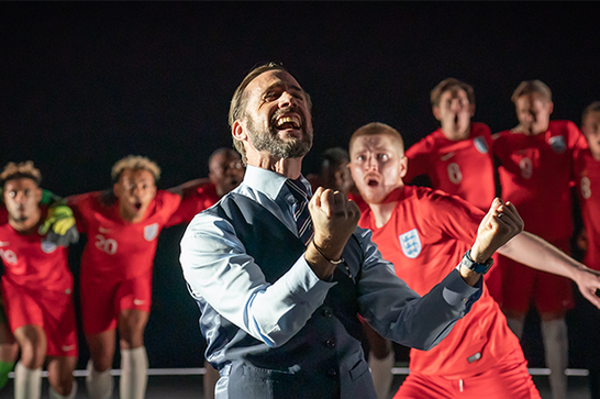 A man in a waistcoat and tie (Joseph Fiennes playing Gareth Southgate) pumps both of his fists in celebration, in front of male England football players watching in the background.