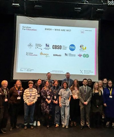 A group of people standing in front of a projection screen, all from the Birmingham Music Education Hub. The screen shows the logo from their respected companies.