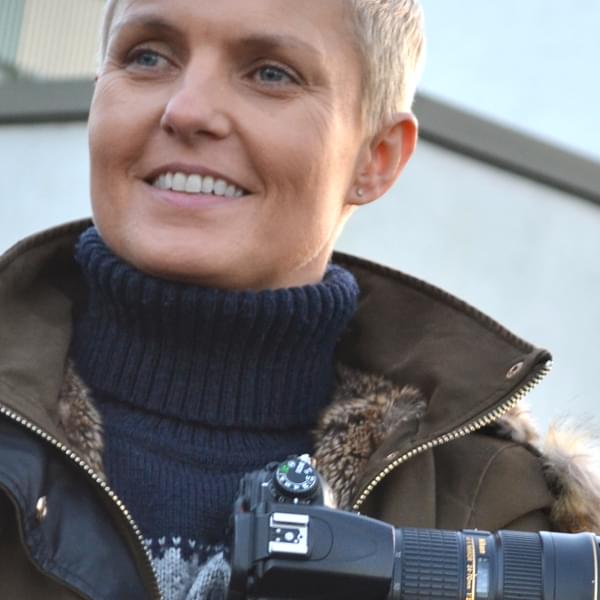 Kate is holding a camera, wearing a khaki fluffy coat. Kate has short blond hair and is smiling into the distance.