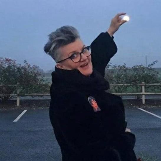 Libby has short grey hair, wearing glasses and a black coat outside, holding her hand up to appear to hold the moon