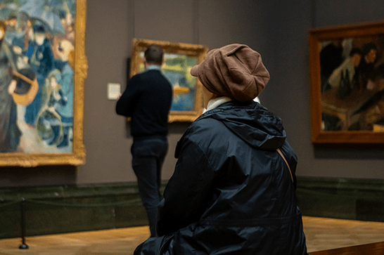 A woman sits and a man stands in a gallery looking at a painting on the wall.