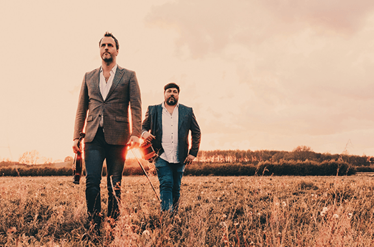 Two men in jackets and holding string instruments walking through a field.