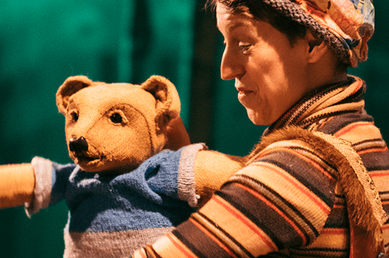 A woman in a striped top and hat holds a teddy bear puppet with a blue jumper.