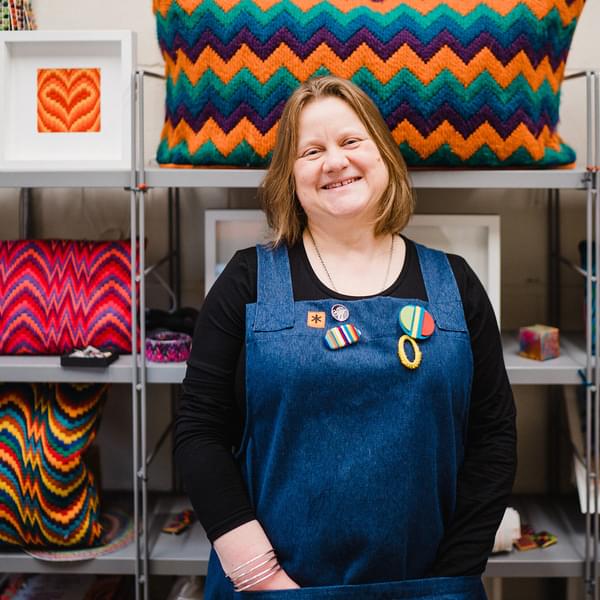 Tina is wearing blue dungarees and has a short brown bob. Tina is stood in front of colourful fabrics.