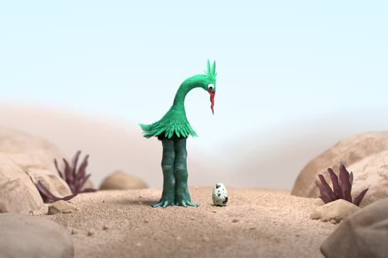 Stood in a desert, a strange green creature looks down at a spotted egg sitting next to it.